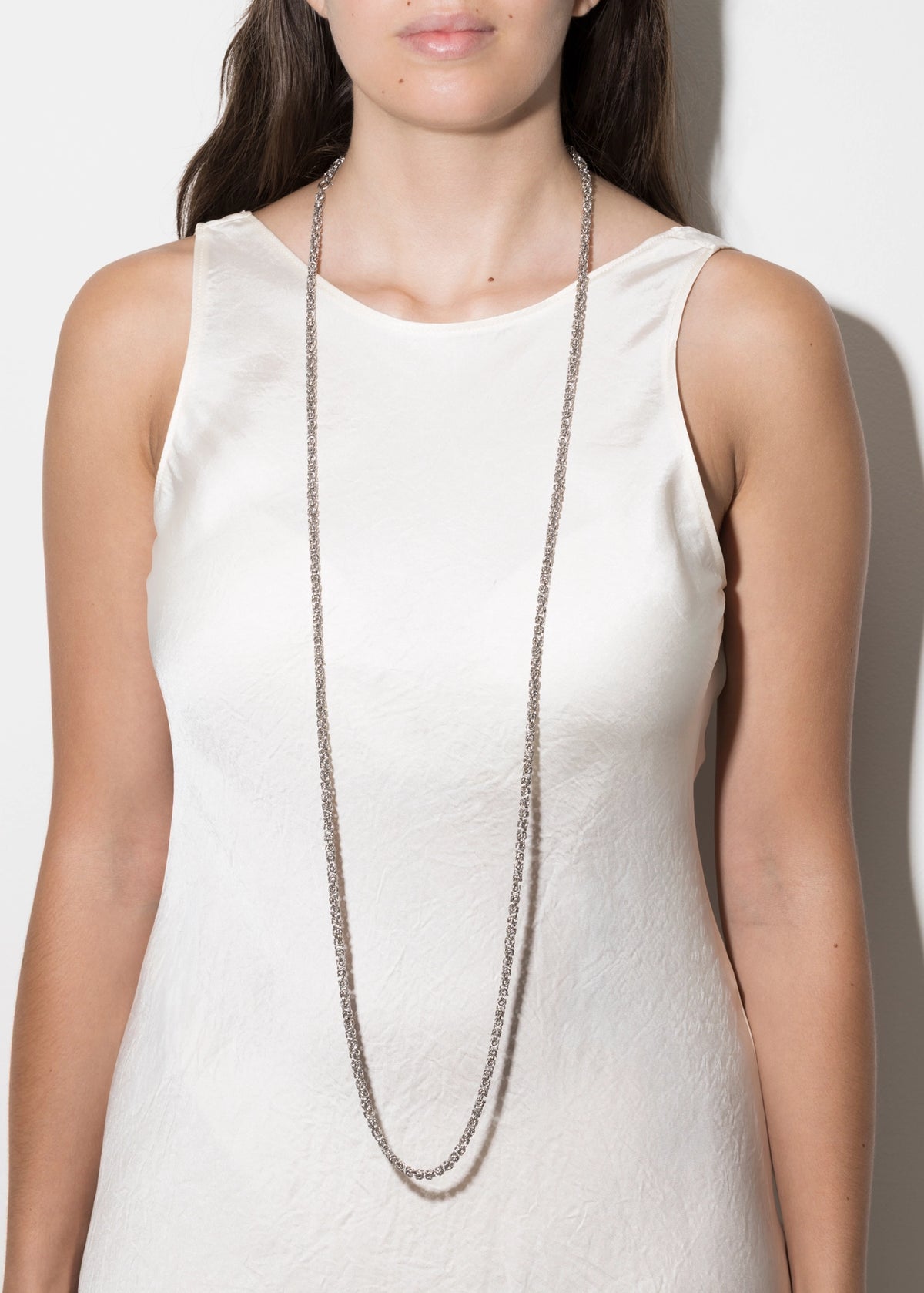 A model wearing a long, double-strand silver chain necklace over a white sleeveless top, shown from the chest up against a plain light background.