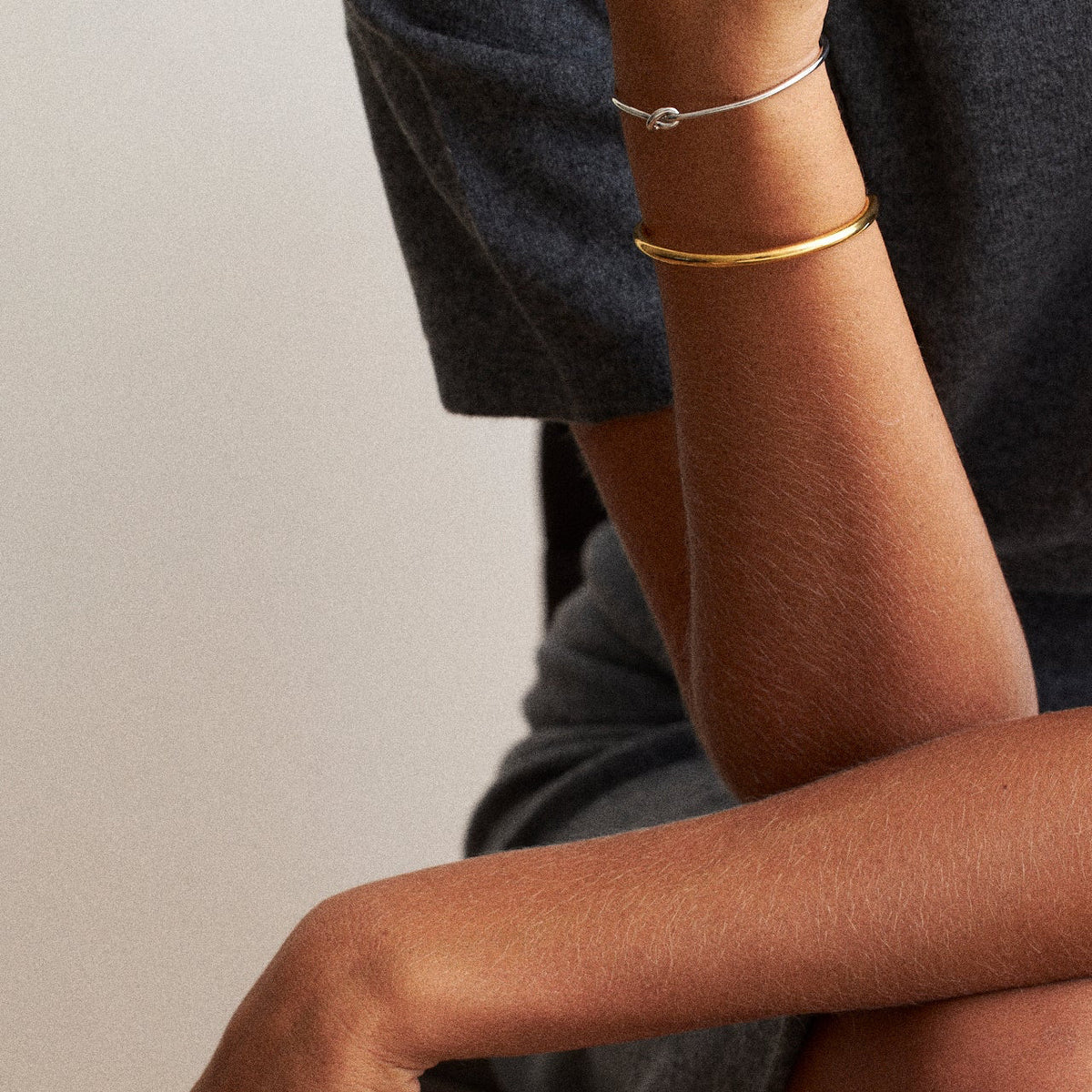 A close-up lifestyle shot of a person wearing two minimalist bangle bracelets, one gold and one silver, on their wrist, paired with a dark gray t-shirt and shorts.