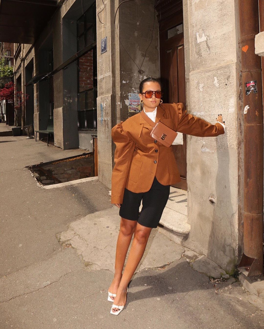 Fashionable woman in oversized brown blazer, black shorts, and white heels posed on street.