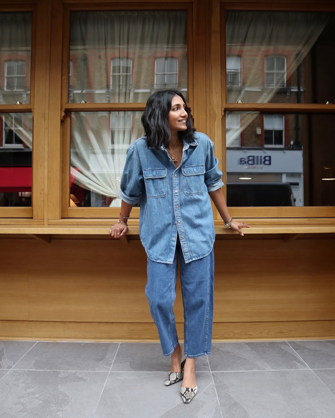 A woman wearing a denim shirt and jeans, smiling in front of a wooden window, showcasing a stylish casual look.