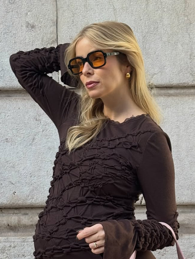 Woman wearing a brown textured dress and sunglasses against a light-colored wall.
