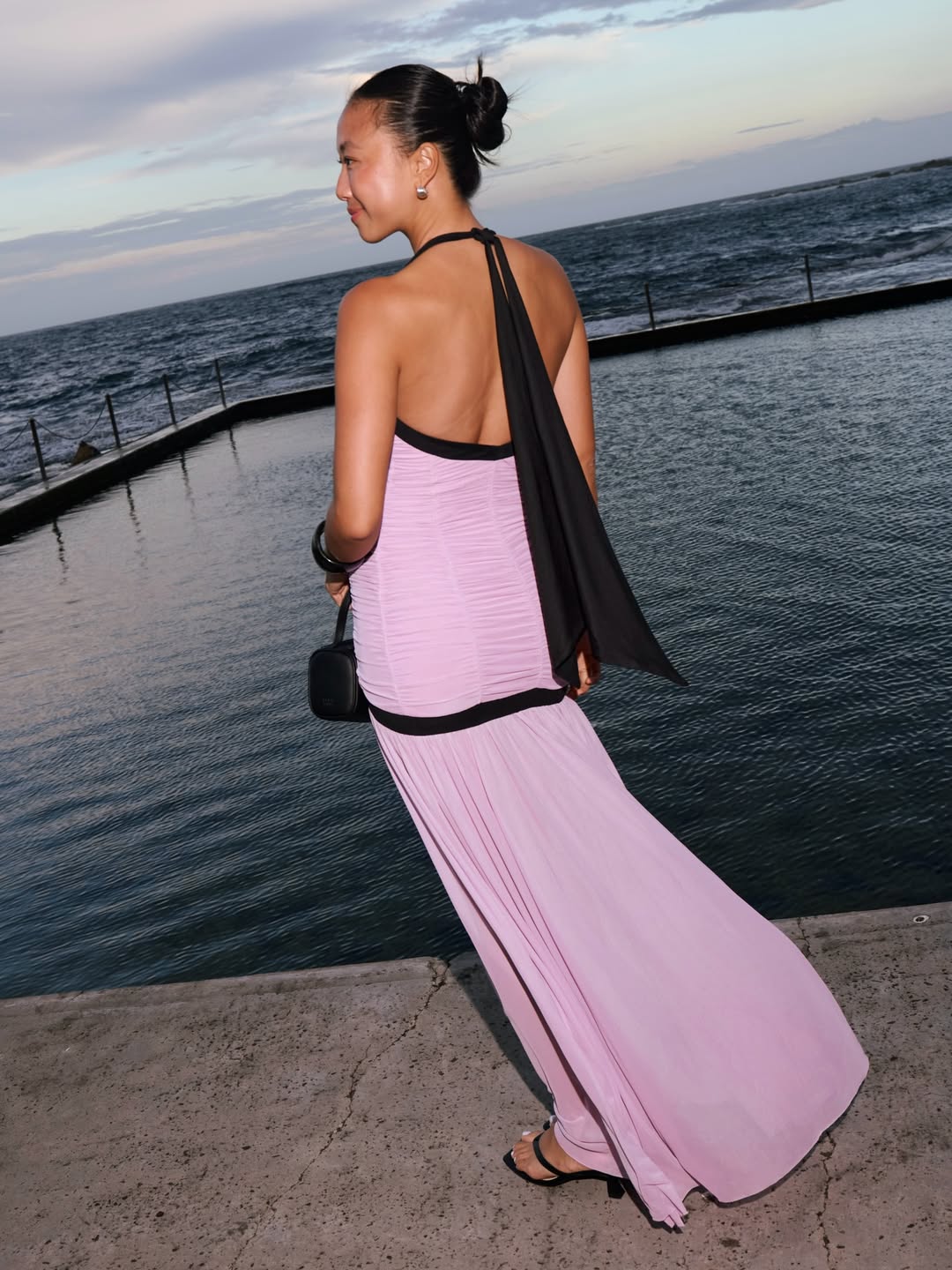 Woman in a pink flowy dress with a black scarf, seen from the back near the water.