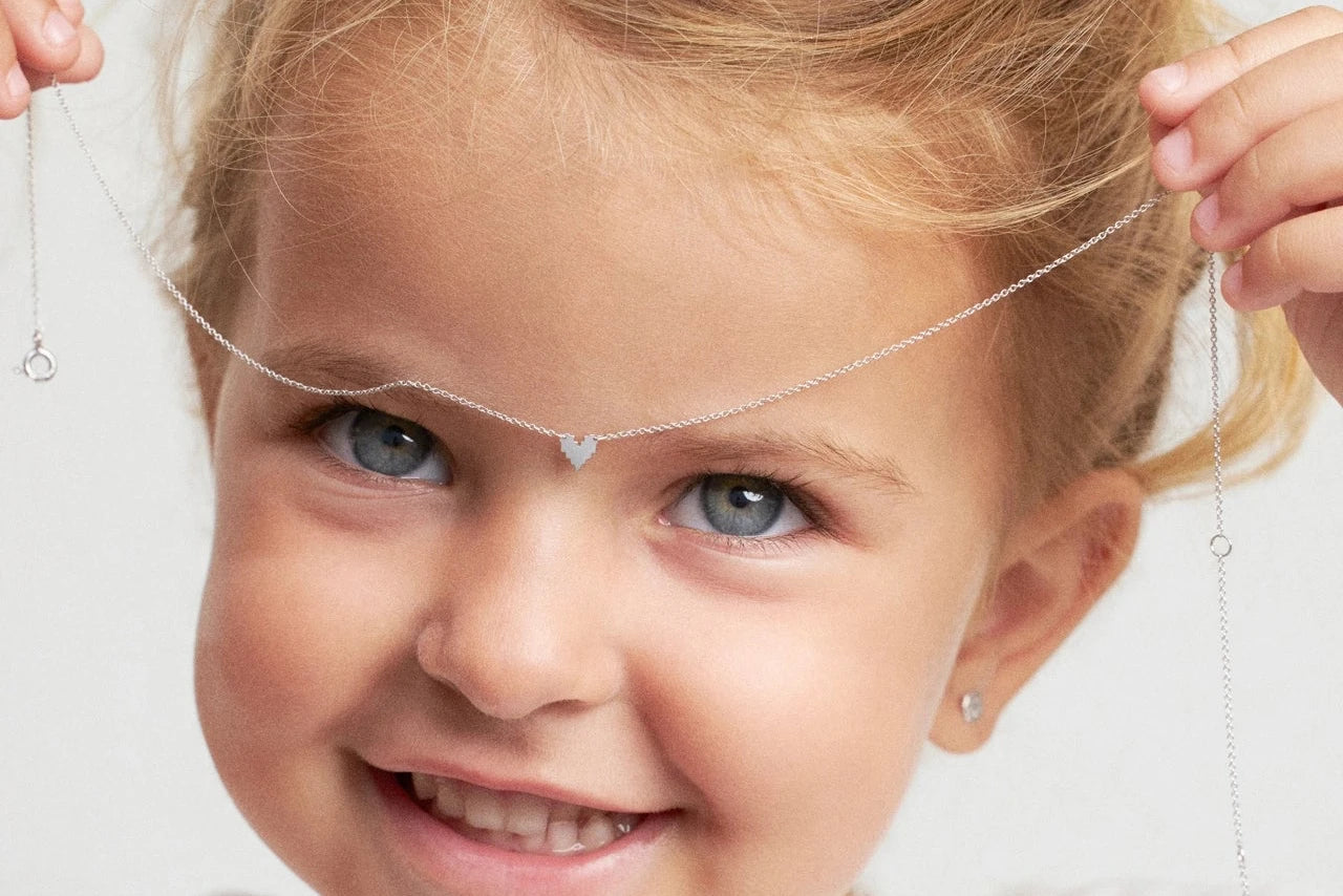 A young child with blonde hair in buns wearing a delicate silver headchain with small dangling heart charms, holding it in place with both hands against a plain light background.
