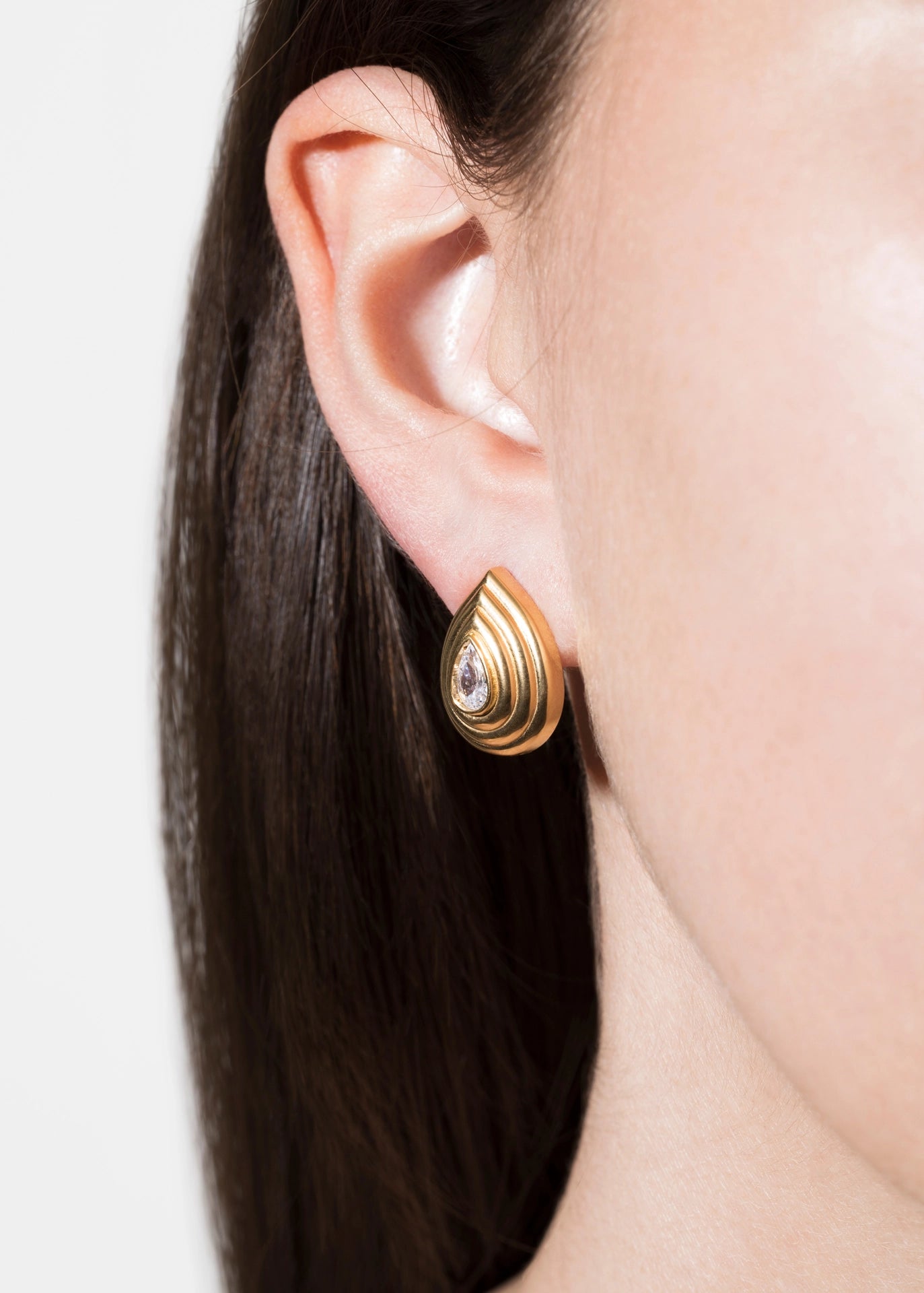 A close-up of a model's ear wearing a gold teardrop-shaped stud earring with concentric ridges and a small clear central stone.