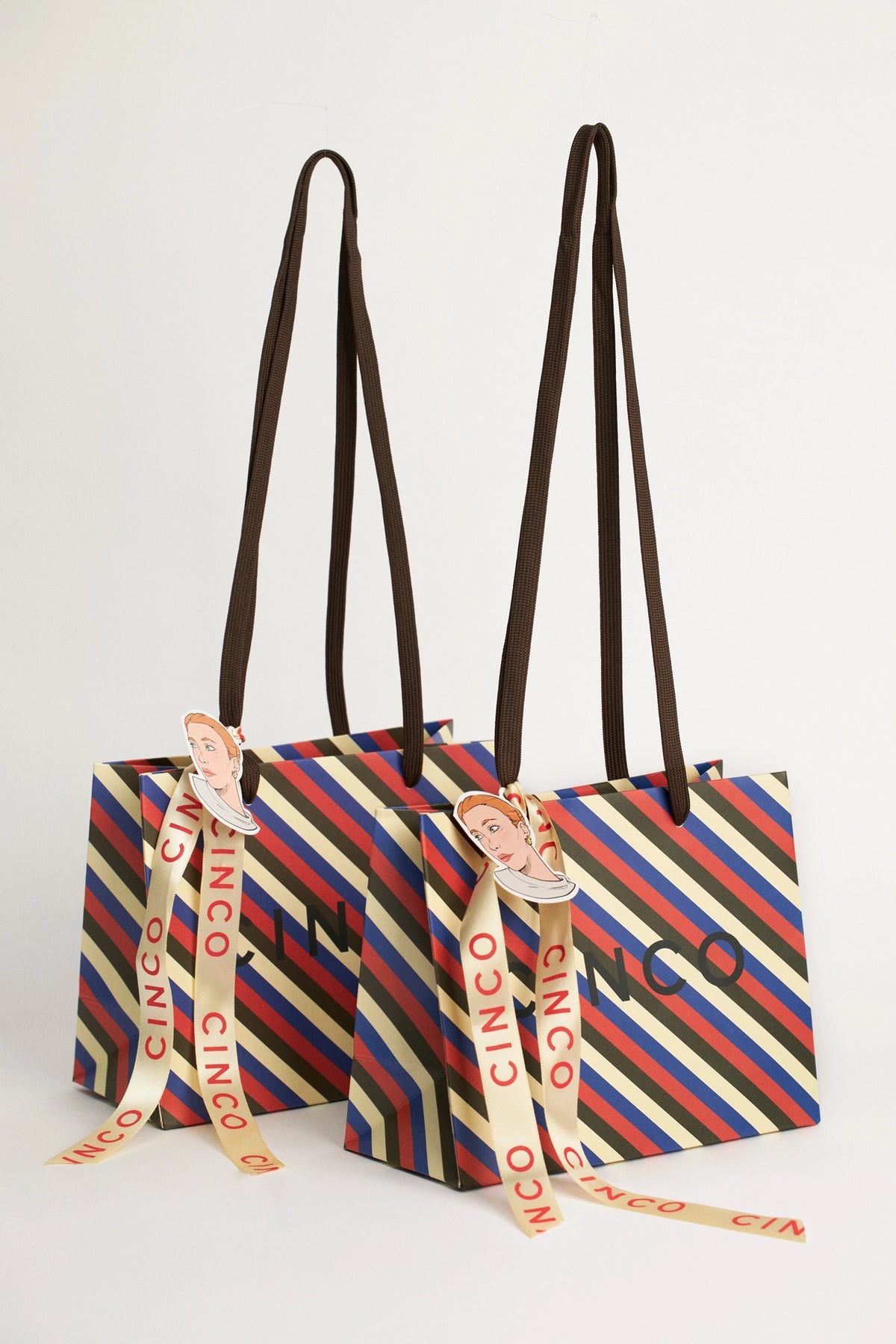 Two Cinco branded paper shopping bags with diagonal multicolor stripes, dark brown shoulder straps, and cream ribbon ties printed with the brand name, shown on a light gray background.