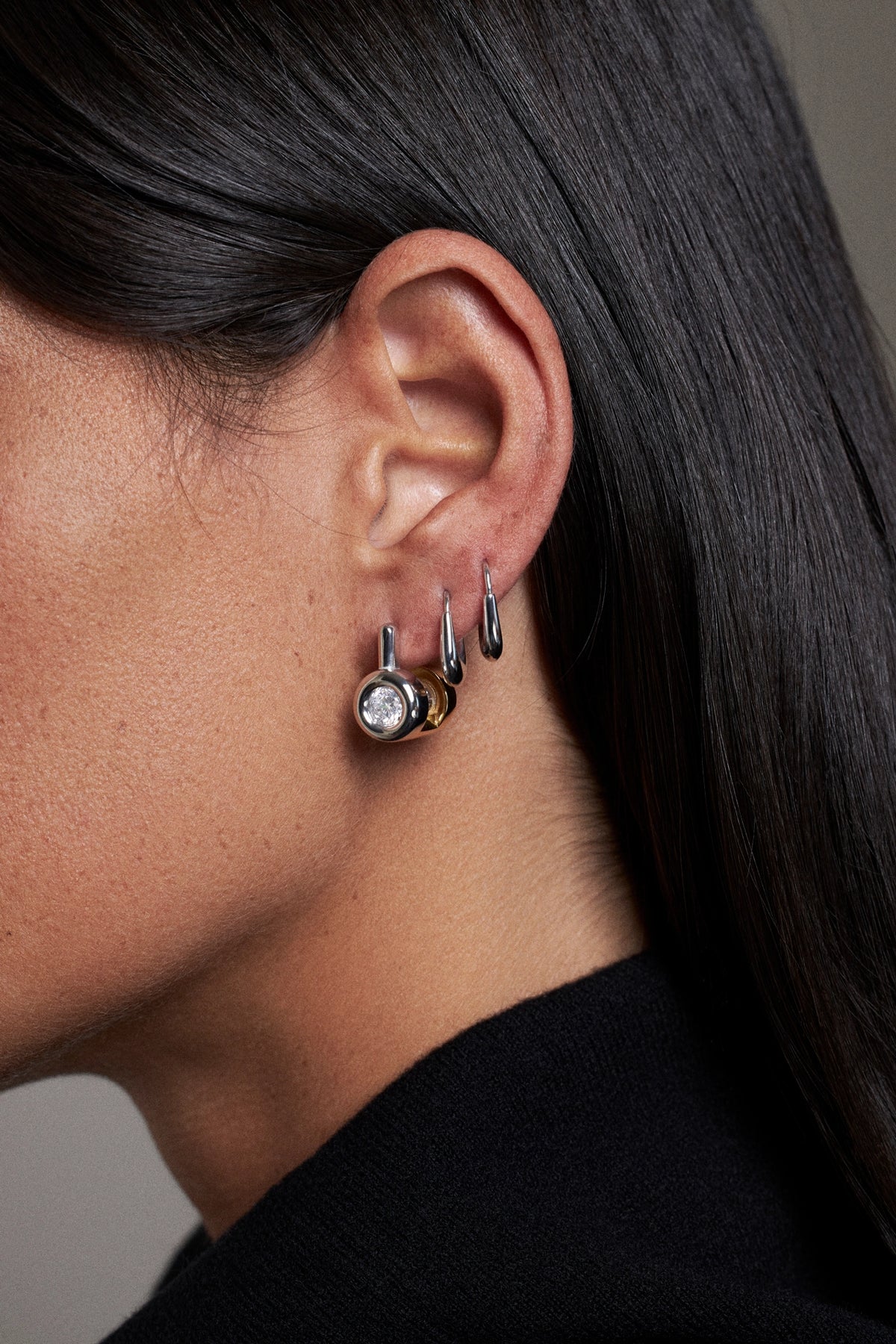 Close-up of a model's ear wearing multiple earrings, including a small silver hoop, a silver hook with a clear round stone, and a gold-toned earring with a dark stone.