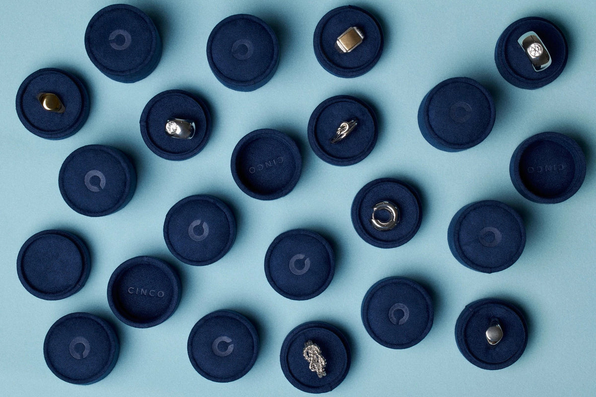 An overhead flat-lay of multiple round dark navy jewelry rings boxes arranged on a light blue background. Several boxes are open, showing small silver and gold jewelry pieces such as rings and earrings.