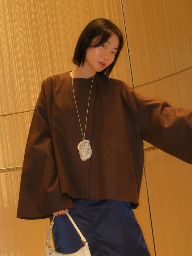 Woman in a brown top and blue skirt holding a white handbag against a wooden wall.